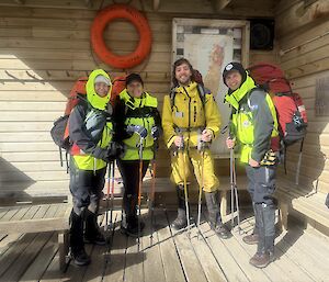Four smiling people wearing outdoor jackets, pants and boots, and wearing backpacks, and carrying walking poles pose for a pre-trip photo.