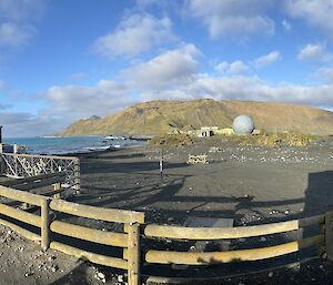 The sun, peeking out from a cloudy sky, illuminates Macquarie Island Station and the green mountainous plateau in the distance.