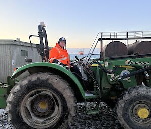 A man wearing a high-vis orange work jacket drives a green tractor - a large orange ship can be seen in the distant background.