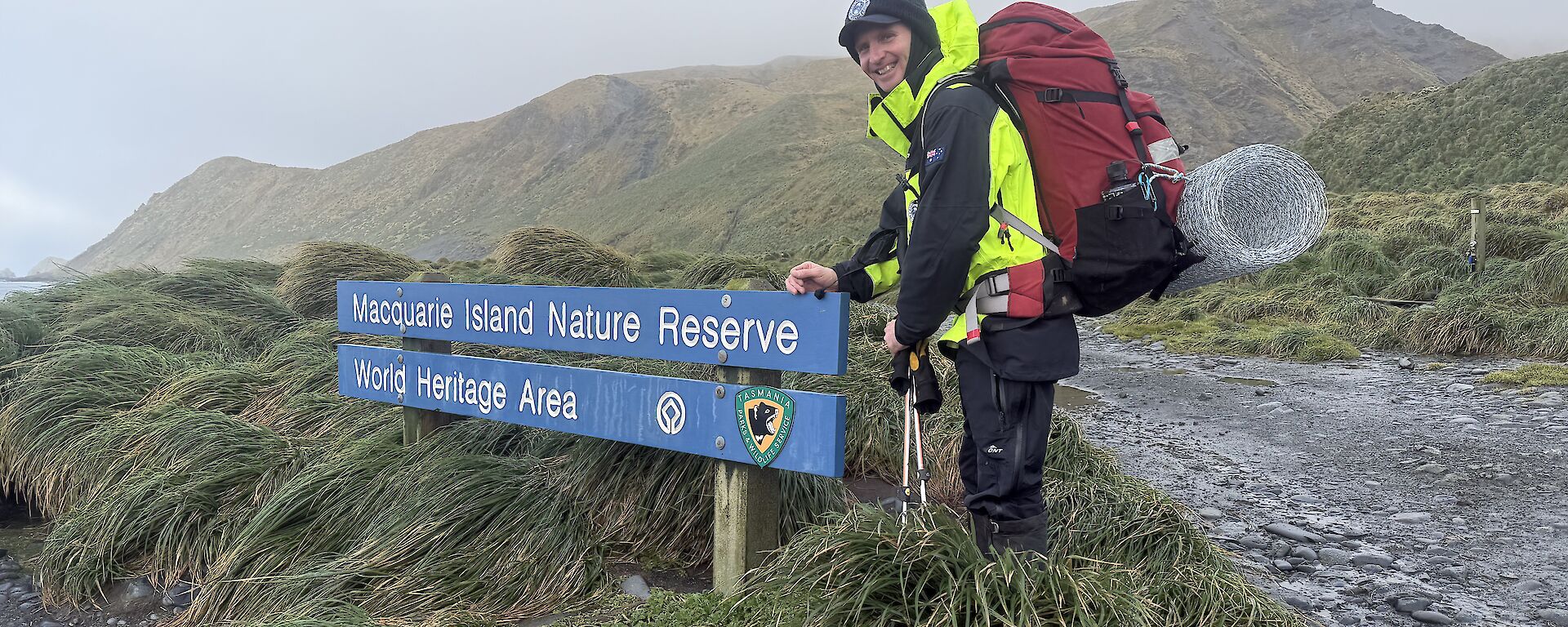 A smiling man wearing outdoor gear and a backpack stands next to a blue sign that says' "Macquarie Island Nature Reserve, World Heritage Area."