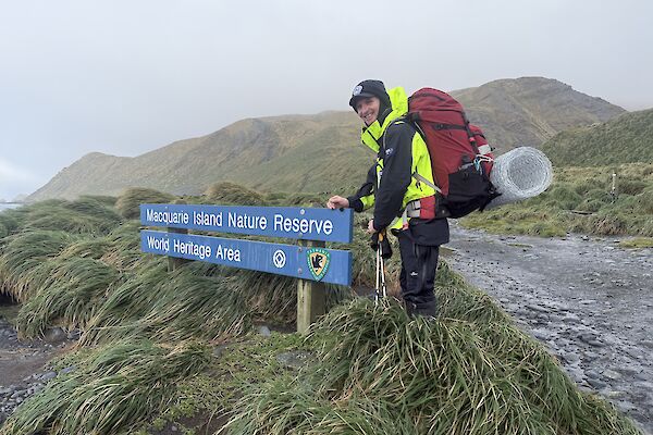 A smiling man wearing outdoor gear and a backpack stands next to a blue sign that says' "Macquarie Island Nature Reserve, World Heritage Area."