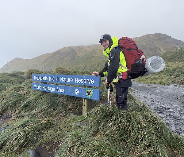 A smiling man wearing outdoor gear and a backpack stands next to a blue sign that says' "Macquarie Island Nature Reserve, World Heritage Area."