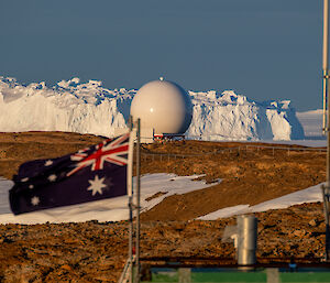The Australian National Flag flies above the barely visible roof of a building with a large white spherical antenna done and a very large iceberg in the distance, under a clear blue sky.