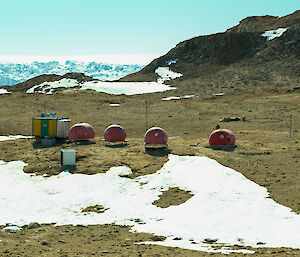 Four round red domes and a green and yellow square hut sit in a rocky landscape with some snow in the foreground