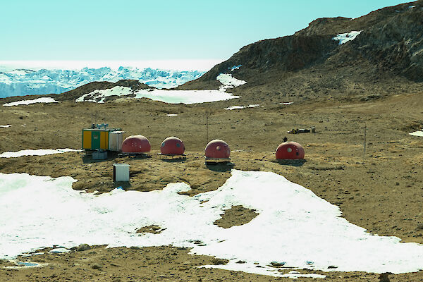 Four round red domes and a green and yellow square hut sit in a rocky landscape with some snow in the foreground