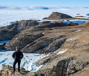 A man wearing black pants and a black puffer jacket stands on a rocky ridge in front of a blue/white frozen glacier