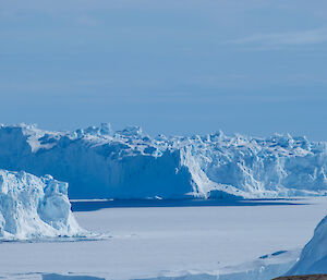 A group of dramatic and craggy blue icebergs rise from a frozen sea with a blue cloudy sky behind them.