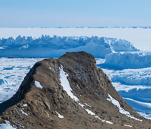 A rocky crag rises from blue/white sea ice, with blue icebergs sitting in the middle distance.