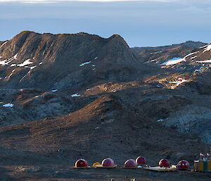 Six red domes, a green and yellow striped hut and a bright yellow tent are nestled in a flat area, with a rocky range rising in the middle distance