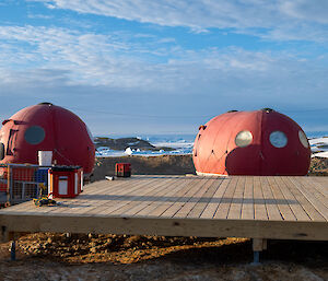 Two Australian National Flags fly over a yellow and green hut, with two red domes sitting behind a large wooden decking platform.
