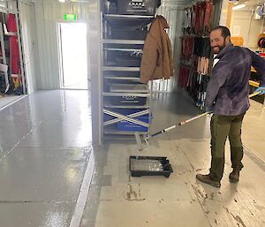 A man in a blue jacket and green pants paints the floor of a storeroom with a roller