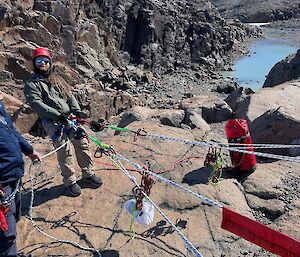 A man wearing green and a harness is collected to a number of ropes and standing near the edge of a rocky cliff