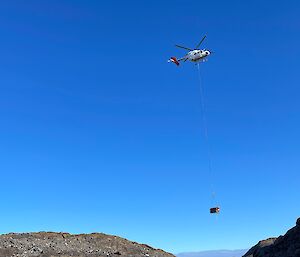 A helicopter with a square load hanging below it flies over a rocky landscape with a bright blue sky