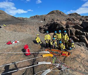 A group of 8 people wearing yellow weather jackets and blue safety helmets stand around a stretcher in a rocky landscape.