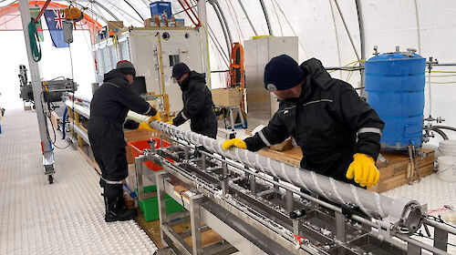 Three people working to remove an ice core from a metal drill barrel, lying on a horizontal bench.