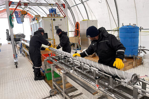 Three people working to remove an ice core from a metal drill barrel, lying on a horizontal bench.