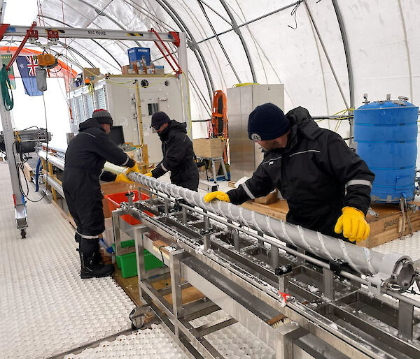 Three people working to remove an ice core from a metal drill barrel, lying on a horizontal bench.