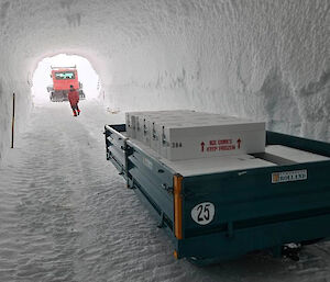 A trailer filled with boxes of ice cores stored inside a tunnel, deep in the ice. At the end of the tunnel is a parked Hagglund vehicle. A person is walking towards the trailer.
