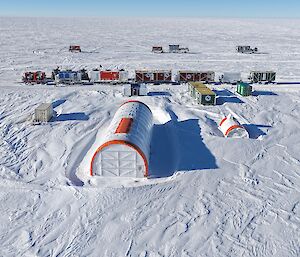 Aeriel view of the drilling campsite showing a line of living and amenities facilities in a series of shipping containers, behind a large semi-circular drilling tent and a smaller tent alongside for storage.