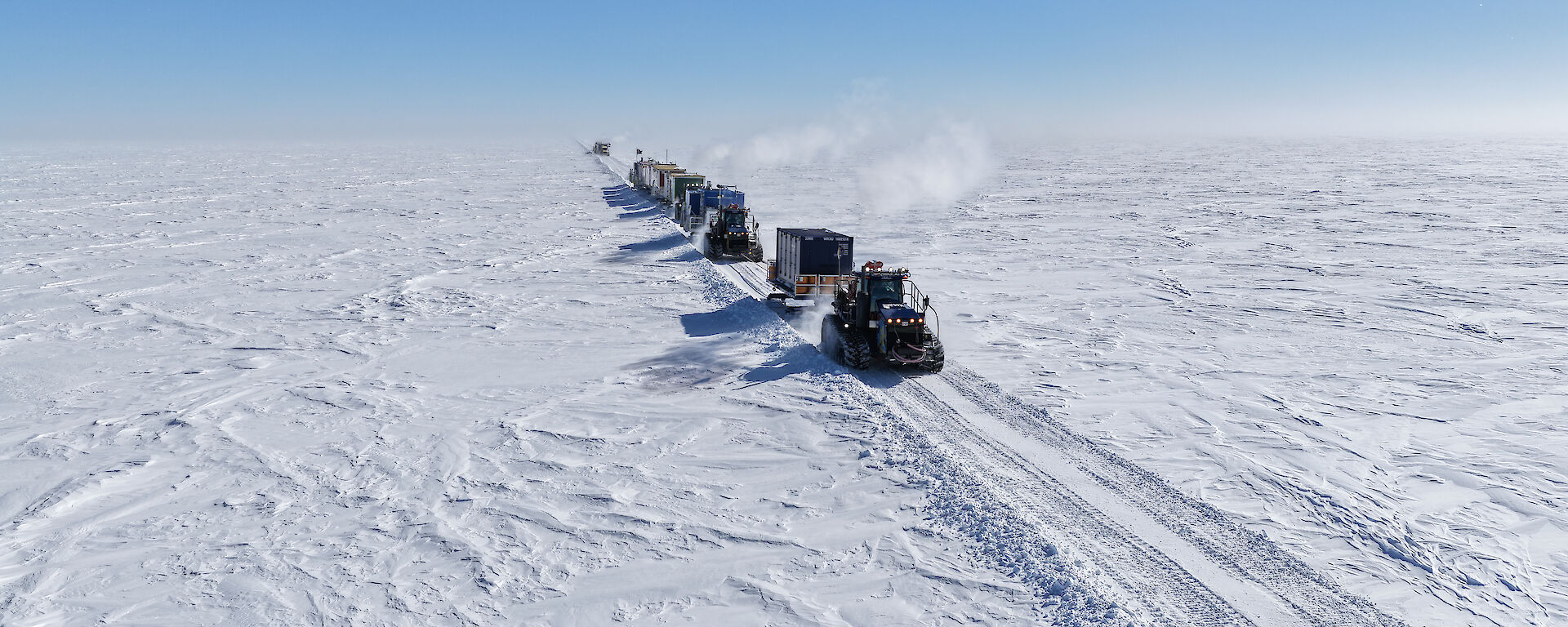 A line of tractors towing sleds carrying shipping containers and equipment on the icy Antarctic plateau.
