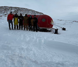 Six expeditioners standing in front of a small hut in Antarctica.