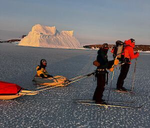Two expeditioners skiing and one sitting down being towed across the ice.