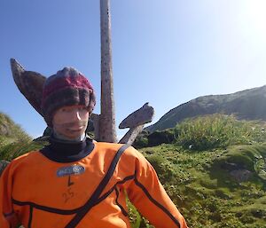 A life sized dummy used for emergency training wears a handmade red and purple beanie while posing against an old rusty anchor set amongst green tussock grass.