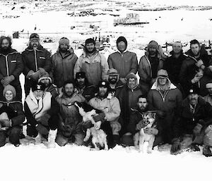 A black and white group photo of expeditioners at Mawson station. Four people kneeling in the front row are nursing huskies.