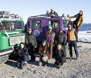 Nine women standing or sitting around two hagglund vehicles, parked on gravel, with ice and a bay in the distance.
