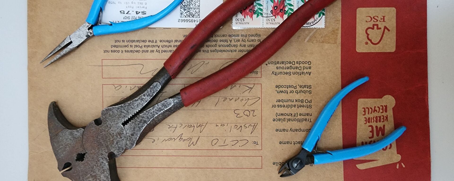 blue handled long-nose pliers, blue handled combination pliers, and a large red handled fencing plier sitting atop an Australia Post envelope addressed to the Communications Technical Officer at Macquarie Island.