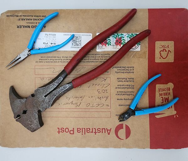 blue handled long-nose pliers, blue handled combination pliers, and a large red handled fencing plier sitting atop an Australia Post envelope addressed to the Communications Technical Officer at Macquarie Island.