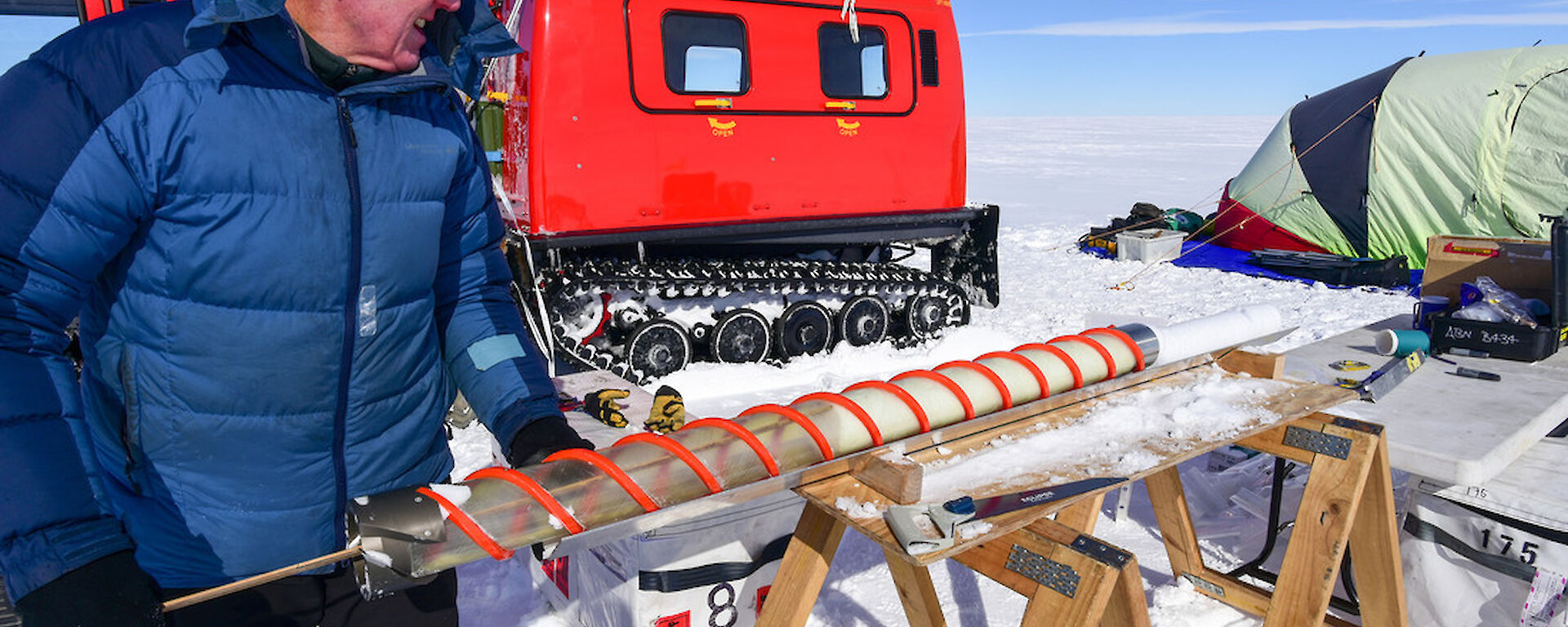 A scientist extracts a firn core from the drill, with a HÃgglunds and dome-shaped tent in the background.
