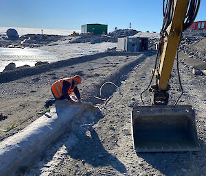 A person in the foreground kneels over the soil bank inspecting the liner, a backhoe bucket in the aisle, with lower Casey pictured in the background