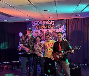 Four people, two with guitars on a stage and standing in front of a band poster showing four seals playing instruments.