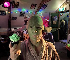 A man with a green painted face, wearing a green beaning and a brown towling dressing gown, holding a small likeness of Yoda from the Star Wars movies.