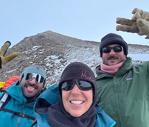 A selfie of three people smiling at the camera with a rocky peak behind.