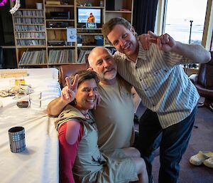 Three people sitting and standing beside a table. Two people are doing V signs with their fingers.