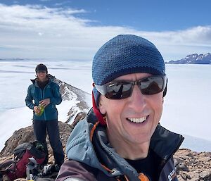 A man in a beanie smiling at the camera, with a woman behind holding a water bottle and smiling at the camera. The two people are standing on a rocky ridge line about the Antarctic ice plateau.