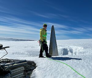 A person clipped in to a green rope standing beside a piece of aircraft tail buried in snow.