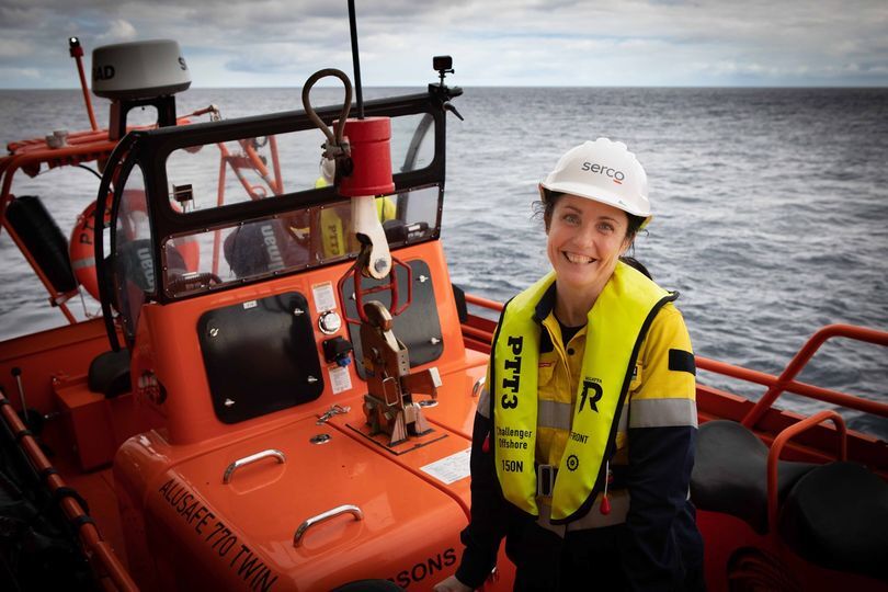 A woman in a hard hat and life jack sits on a barge at the side of a ship smiling to camera